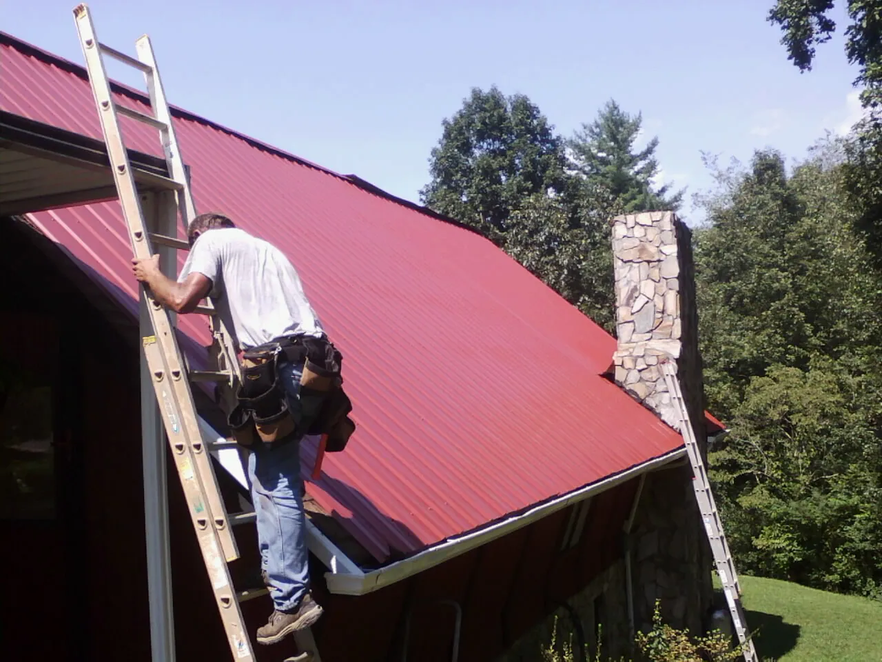 Roofer on a ladder beside a red metal roof and stone chimney — Soffit & Fascia Repair by Roofing Brothers in Ariel